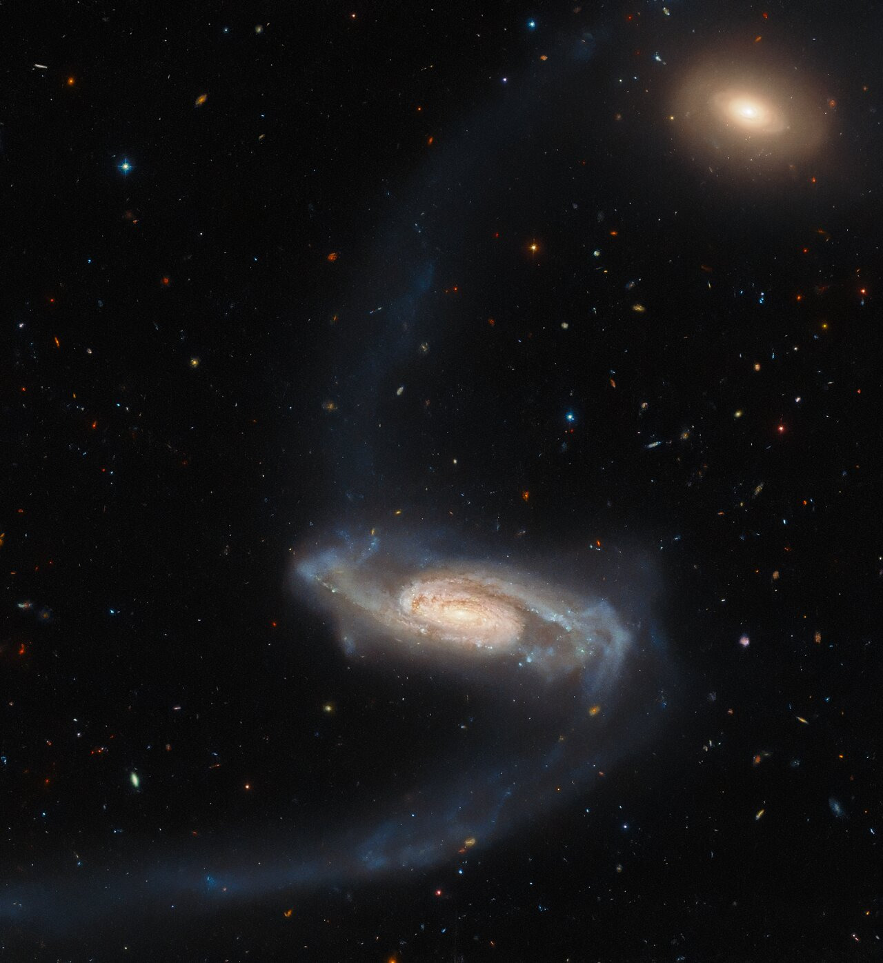 A spiral galaxy. It has a bright core with patches of dark dust, and fuzzier, dimmer spiral arms in cooler colours, with spots of bright blue. Long, faint tidal streams stretch from the galaxy’s arms: one up to the top of the frame, one curving down to the bottom-left corner. In the top-right there is a smaller, orange elliptical galaxy. The background is studded with many tiny stars and galaxies.